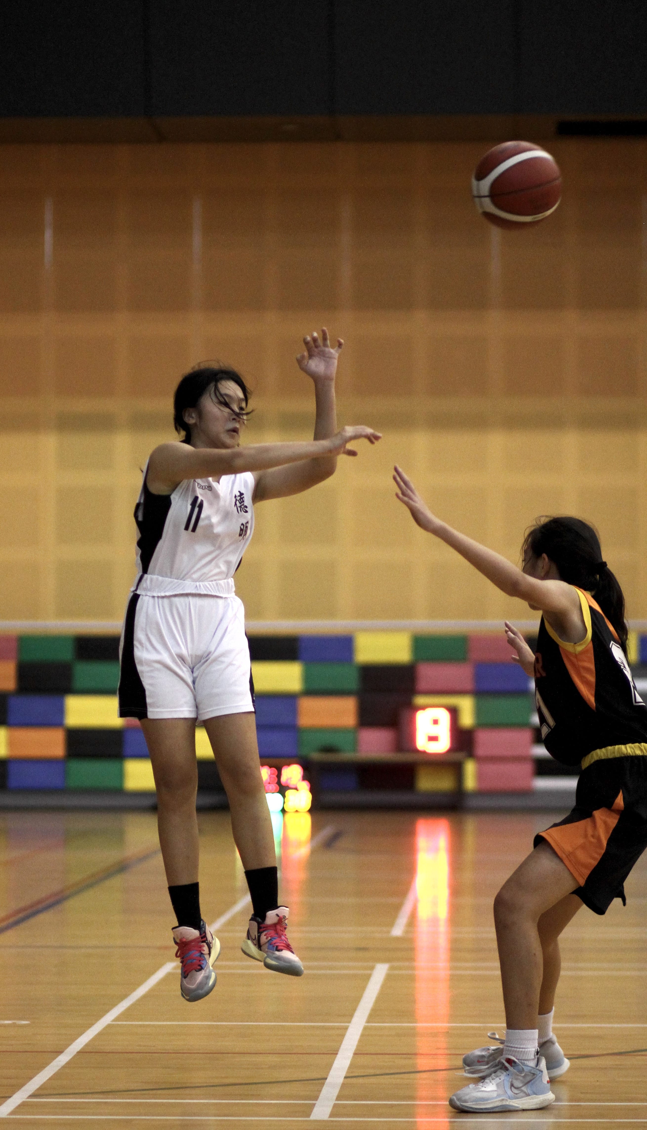 Player in white jersey number 11 jumps to pass a basketball while a defender in black and orange reaches up.